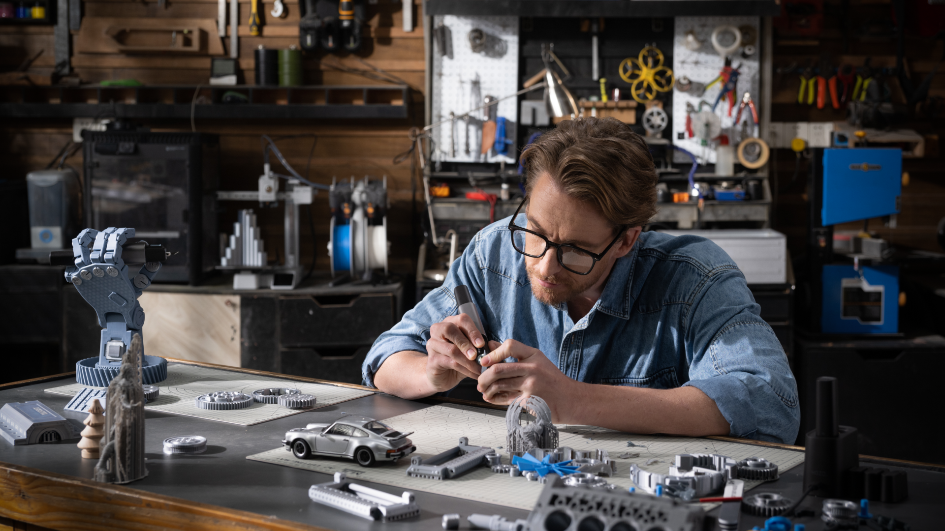A man in a garage workshop carefully post-processing 3D printed models, including a mechanical hand and a toy car.