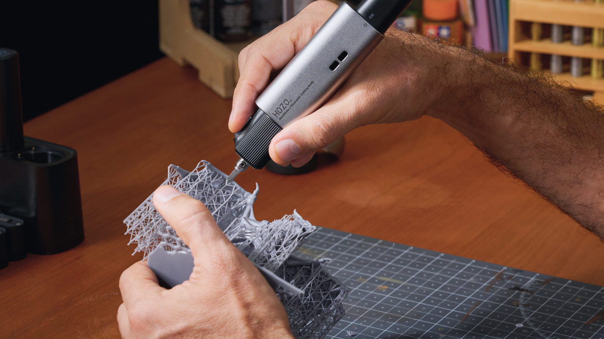 A person uses a HOZO ultrasonic cutting knife to remove dense supports from a gray resin miniature above a cutting mat on a workbench.