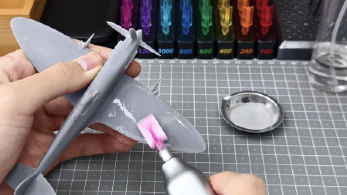 A person sanding a gray plastic aircraft model with a pink polishing tool on a cutting mat, with sandpaper sets and a metal dish nearby.