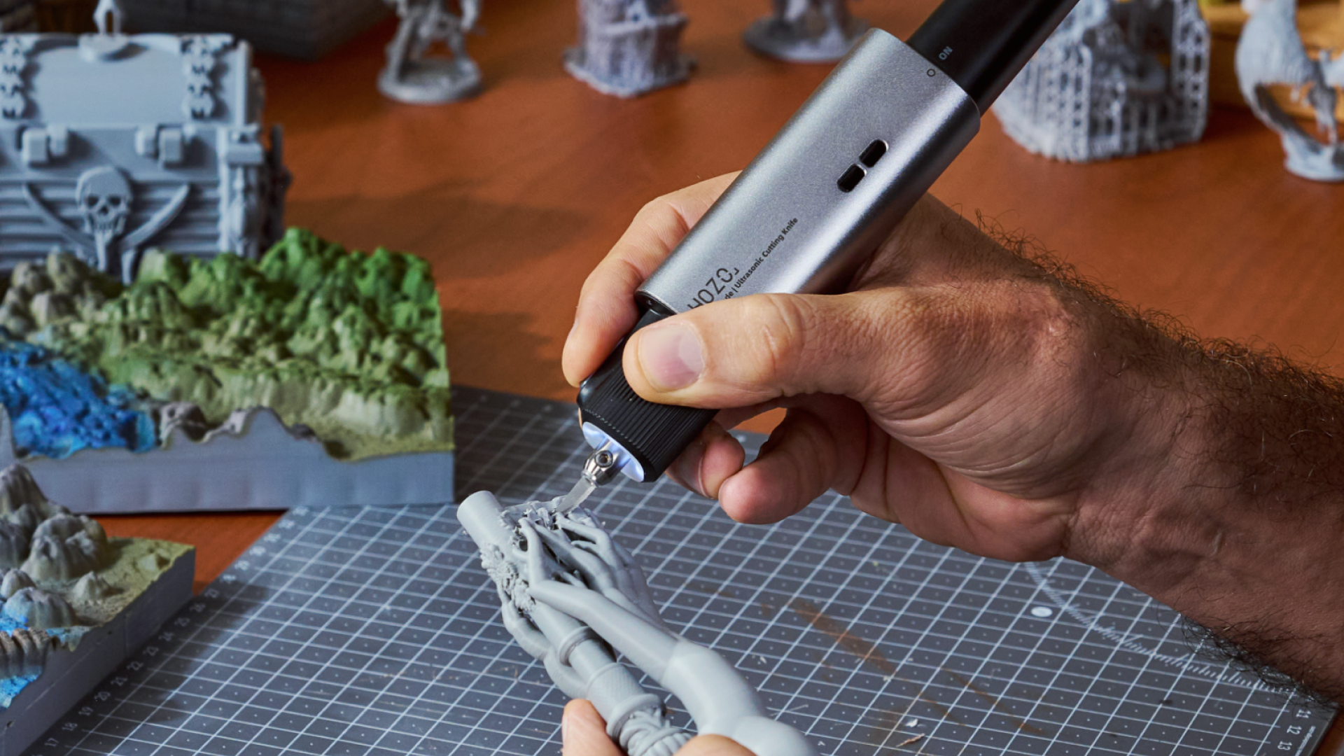 A close-up view of a user's hand guiding the NeoBlade ultrasonic hobby knife to carefully detach complex support structures from a highly detailed grey 3D printed miniature.