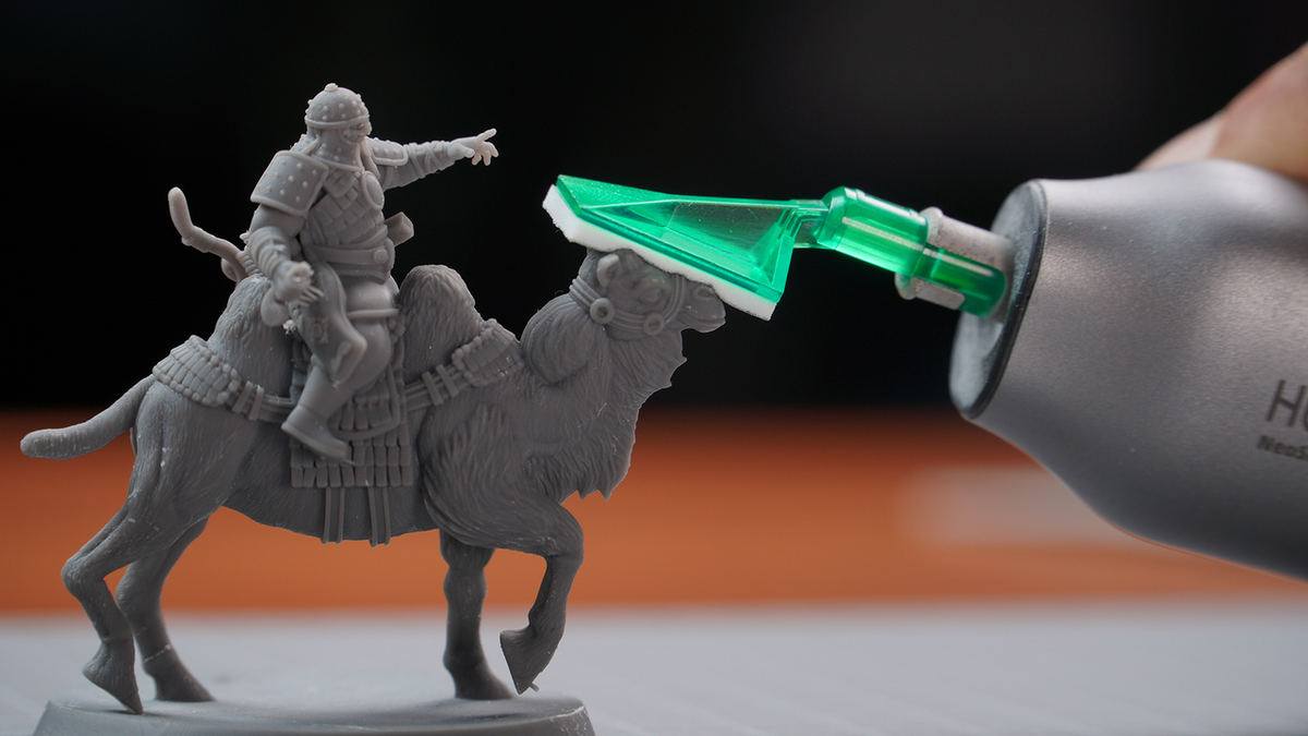 Hand with a green-tipped tool touching the face of a grey camel rider figurine.