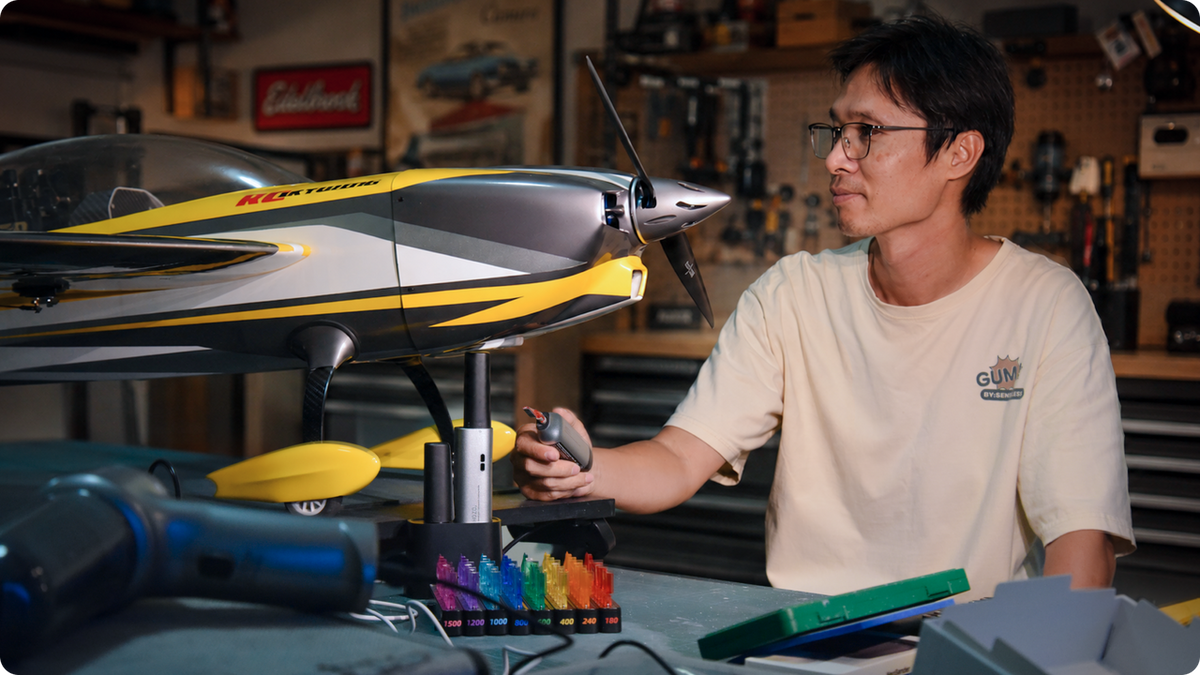 RC pilot Shark sits at his workbench, preparing to modify a yellow balsa-wood RC plane into a custom FPV aircraft using precision crafting tools.