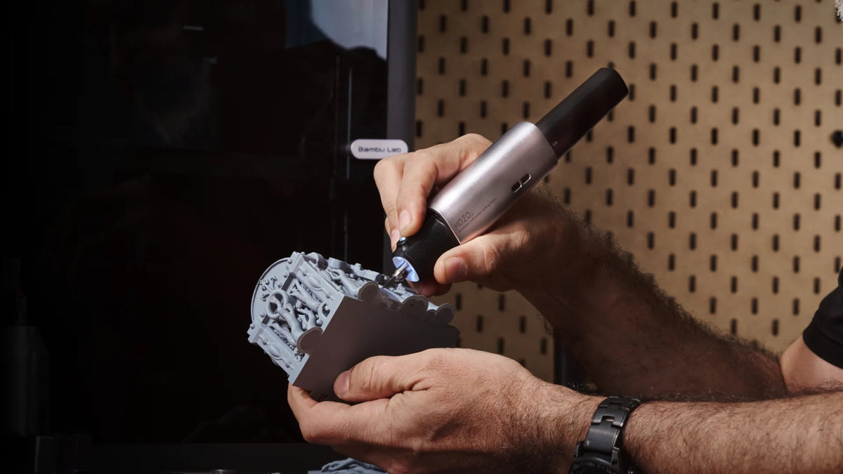 Hands holding an ultrasonic cutter knife, applying it to a small silver cylinder on a black block.